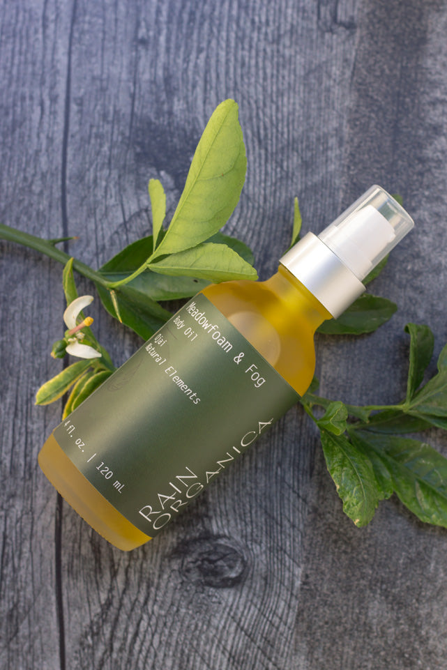 Rain Organica Ojai body oil (4 ounce) lying on a weathered grey wooden table with a sprig of orange tree with orange blossom underneath the bottle